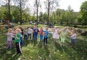 Dzieci w luźnej grupie stoją na trawie w ogrodzie przedszkolnym. Patrzą w jednym kierunku. Niektóre z nich podnoszą ręce do góry.